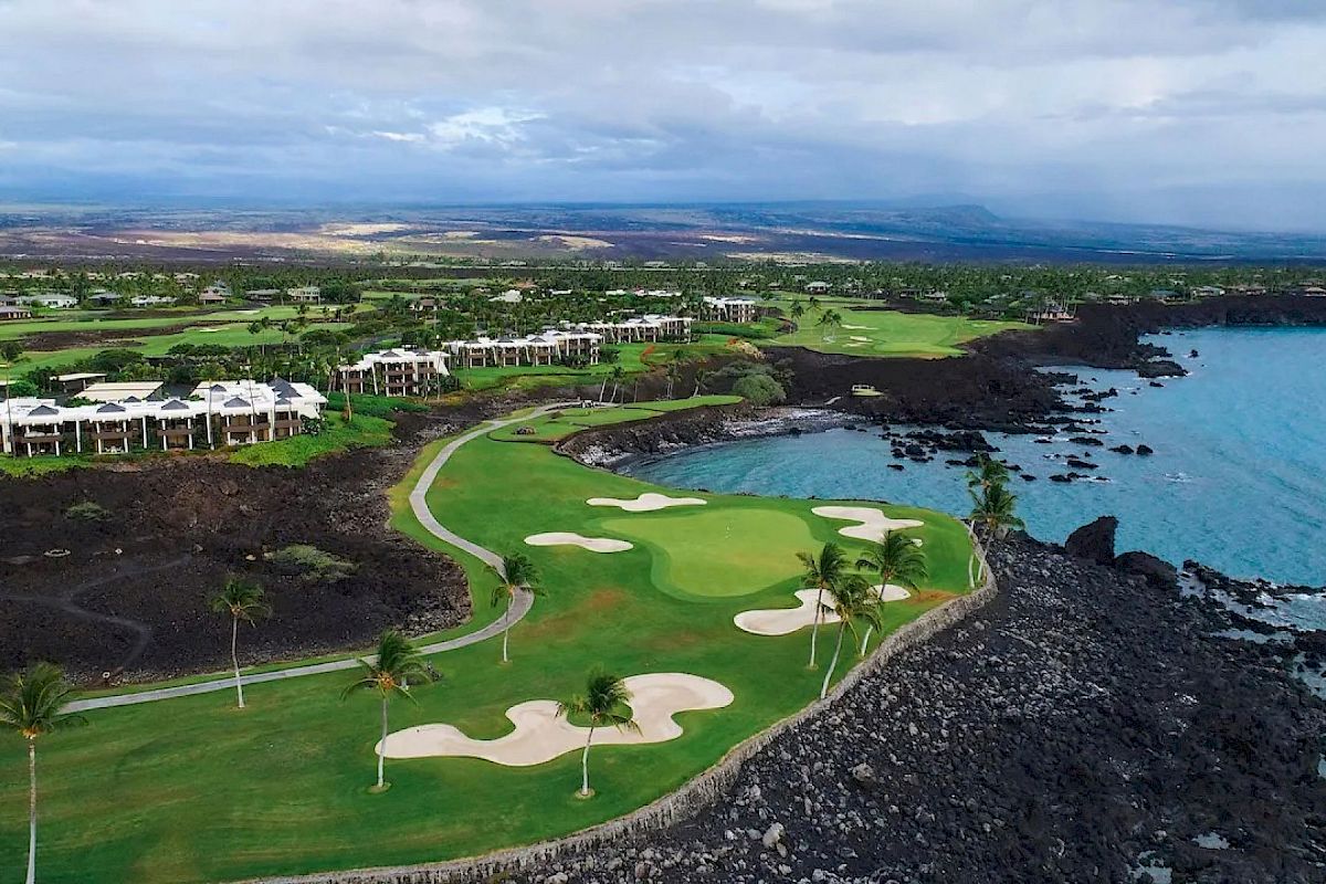 A coastal golf course surrounded by lush greenery, buildings, and rocky shores with an ocean view in the distance under a partly cloudy sky.