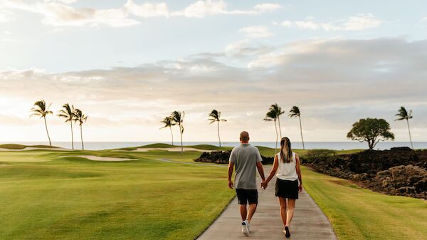 A couple is walking hand in hand on a pathway, surrounded by lush green grass, palm trees, and a scenic ocean view in the background, at sunset.