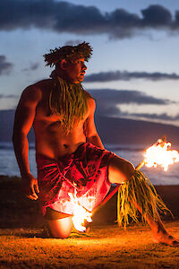 A person performs a fire dance on a beach at sunset, wearing traditional attire with flames in each hand and the background of the ocean.