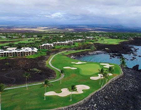 The image shows a scenic golf course by the ocean, with green fairways, sand bunkers, palm trees, and residential buildings in the background.