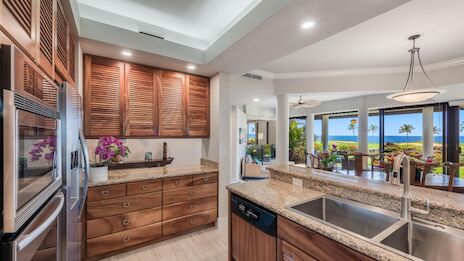 A modern kitchen with wooden cabinets, granite countertops, stainless steel appliances, and a view of a dining area leading to the ocean.