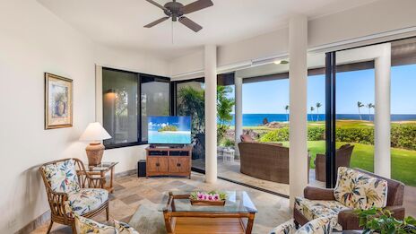 A bright coastal living room with ceiling fan, tropical-patterned seating, large glass doors opening to a veranda and ocean view, sunny and inviting.