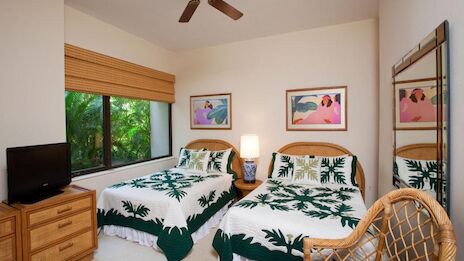 Two beds with matching tropical bedding, a small dresser with a TV, a window with a blind, two framed palm prints, and a wicker chair in a bright, tropical hotel-style room.