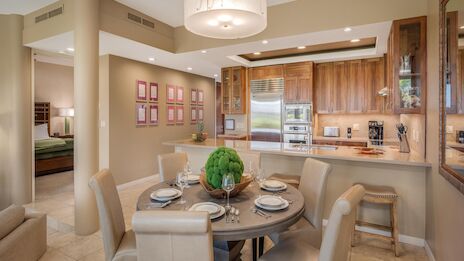 A modern open-concept kitchen and dining area with a round table set for four, cream chairs, wooden cabinets, and stainless steel appliances in warm beige tones.