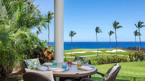A cozy patio with a round table and chairs, tropical plants, palm trees, and a sunny view of a golf course and the blue ocean beyond, perfect for a relaxing meal.