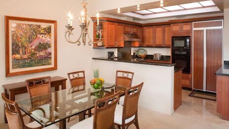 A modern kitchen and dining area with wooden cabinets, stainless fridge, glass-top dining table, chandelier, and framed wall art.