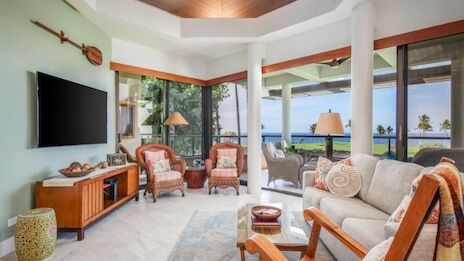 A bright coastal living room with a wall-mounted TV, wicker chairs, a sofa, plants, and large glass doors opening to a sunny ocean view, inviting and airy.