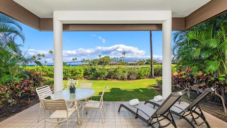 A covered patio with tiled floor, outdoor dining table and chairs, two lounge chairs, lush tropical plants, and a sunny lawn with palm trees beyond the frame.