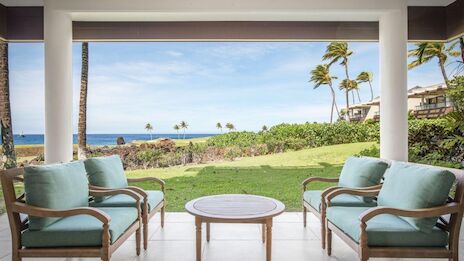 An outdoor patio with four cushioned chairs and a round table, overlooking a grassy area with palm trees and an ocean view.