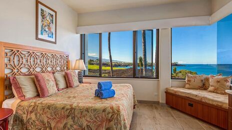 A bright hotel room with a king bed, patterned bedding, a wooden headboard, a bench by three windows, and a view of palm trees and the ocean.