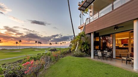 A sunny beachfront house with a grassy yard, glass-walled patio, palm trees, and a stunning ocean view at sunset.
