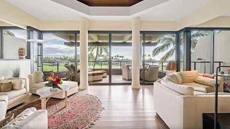 A bright living room with large windows, tropical views, cozy sofas, a round rug, and a balcony with outdoor seating.