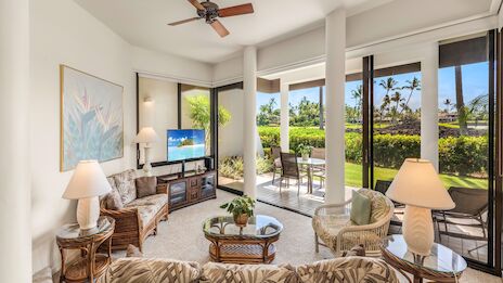 A bright tropical living room with beige seating, glass coffee table, TV stand, lamps, ceiling fan, and a balcony overlooking greenery and blue skies.