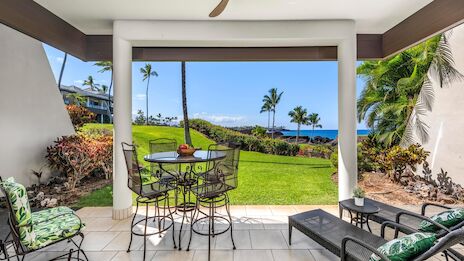 A covered patio with a metal table and chairs, a lounge chair, palm trees, a sunny lawn, and a view of the ocean.