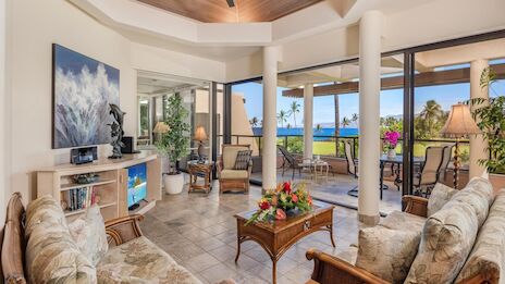 A bright, airy living room with beige floral sofas, a wooden coffee table, TV stand, potted plants, and large glass doors opening to a patio and ocean view.