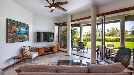 A modern living room with a sofa, TV, artwork, and large windows offering a view of a sunny garden and patio furniture outside.