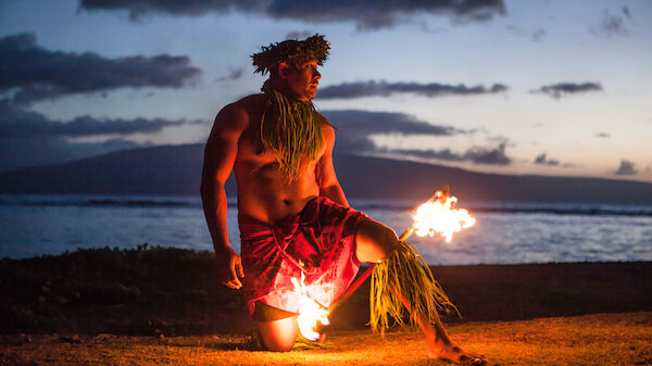 A person is performing a traditional fire dance by the ocean at sunset, wearing cultural attire with flaming torches in hand.