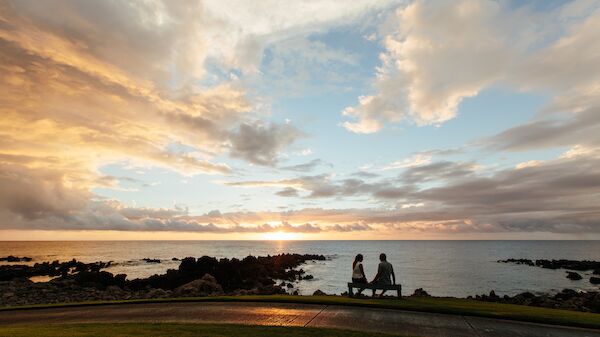 A couple sits on a bench by the ocean at sunset, surrounded by rocks and with a colorful sky, creating a romantic atmosphere.