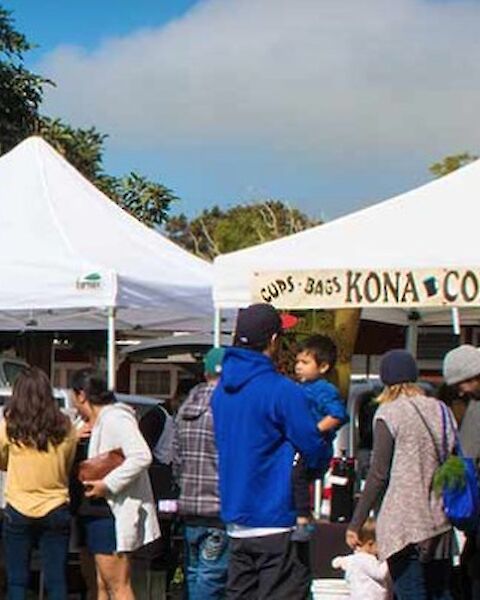 People are gathered around tents at an outdoor market, with one tent labeled "Kona Coffee Espresso."