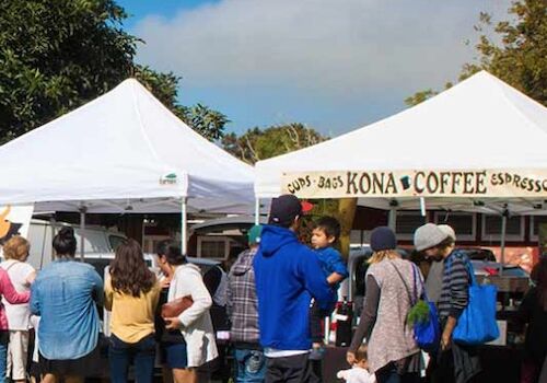 People are gathered around tents at an outdoor market, with one tent labeled "Kona Coffee Espresso."