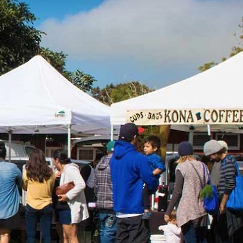 People are gathered around tents at an outdoor market, with one tent labeled "Kona Coffee Espresso."