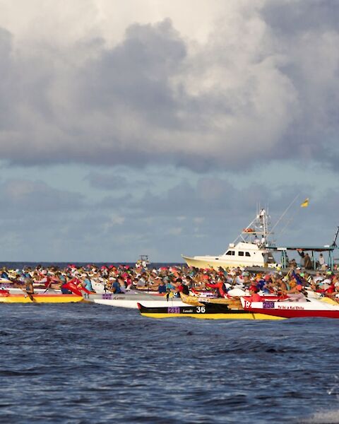 The image shows a canoe race with multiple teams on the water, accompanied by a boat under a cloudy sky.