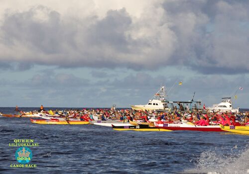 The image shows a canoe race with multiple teams on the water, accompanied by a boat under a cloudy sky.