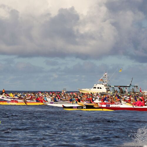 The image shows a canoe race with multiple teams on the water, accompanied by a boat under a cloudy sky.