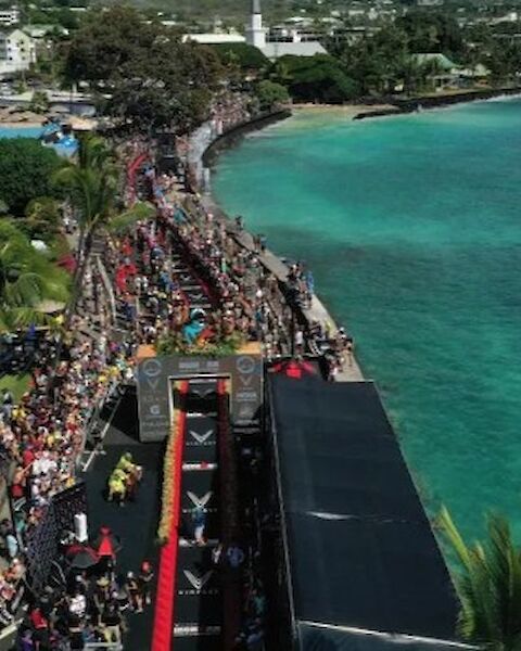 Aerial view of a coastal race event with a crowd, banners, a finish line structure, and clear ocean waters on the right.