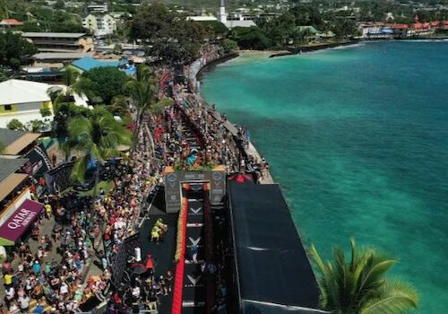 Aerial view of a coastal race event with a crowd, banners, a finish line structure, and clear ocean waters on the right.