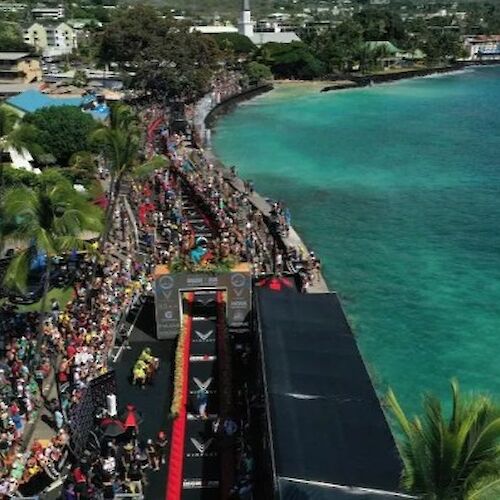 Aerial view of a coastal race event with a crowd, banners, a finish line structure, and clear ocean waters on the right.