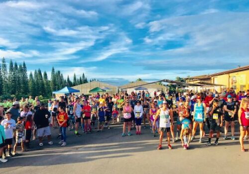 A crowd of people, including runners, is gathered outdoors under a blue sky with tents and trees in the background.