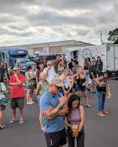 A crowd of people gathered at a food truck event in an outdoor setting with several food trucks visible in the background.
