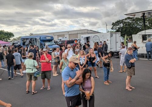 A crowd of people gathered at a food truck event in an outdoor setting with several food trucks visible in the background.