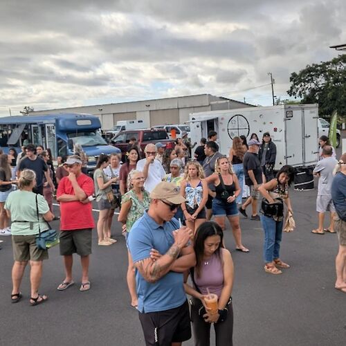 A crowd of people gathered at a food truck event in an outdoor setting with several food trucks visible in the background.
