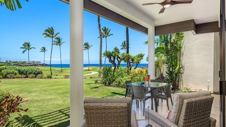 A sunny beachfront patio with wicker seating, a dining table, palm trees, and a view of grassy lawn leading to the ocean, under a ceiling fan.