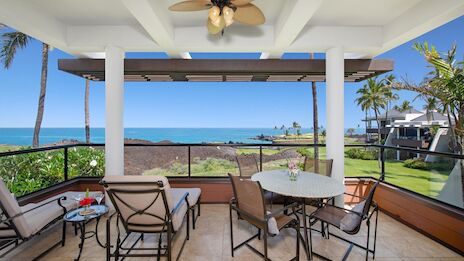 A beachfront patio with a dining table and chairs, ceiling fan, palm trees, and a view of the blue ocean and clear sky.