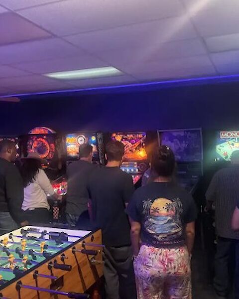 A lively arcade with people playing retro pinball and arcade games; neon lights, foosball table, and a busy, colorful gaming room.