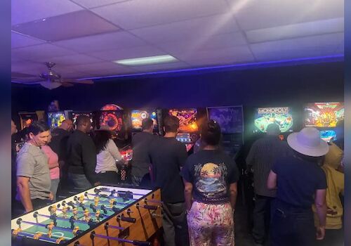 A lively arcade with people playing retro pinball and arcade games; neon lights, foosball table, and a busy, colorful gaming room.