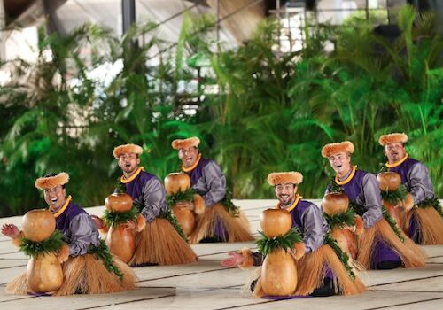 A group of small monkey figurines dressed in purple tops and brown skirts are arranged in a line on a tiled surface, with green plants in the background.