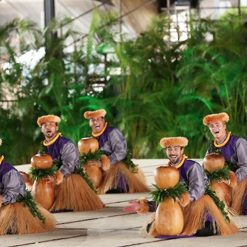 A group of small monkey figurines dressed in purple tops and brown skirts are arranged in a line on a tiled surface, with green plants in the background.
