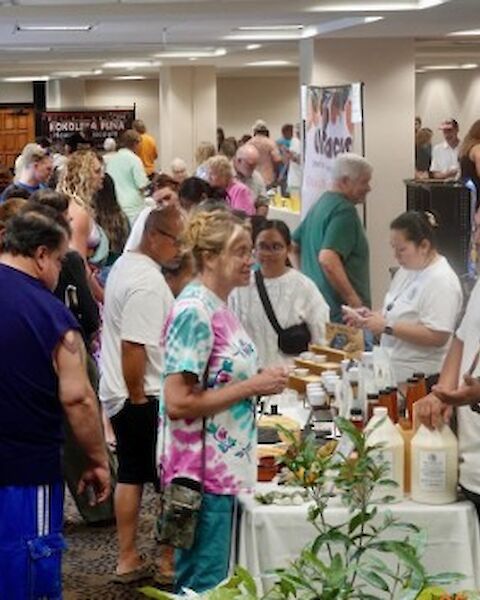 A crowded indoor fair or market with many people browsing stalls and sampling items; enthusiastic vendors chat with attendees.