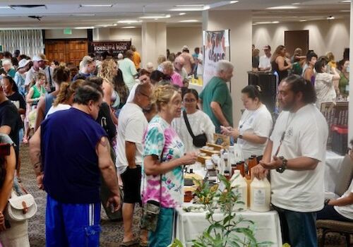 A crowded indoor fair or market with many people browsing stalls and sampling items; enthusiastic vendors chat with attendees.
