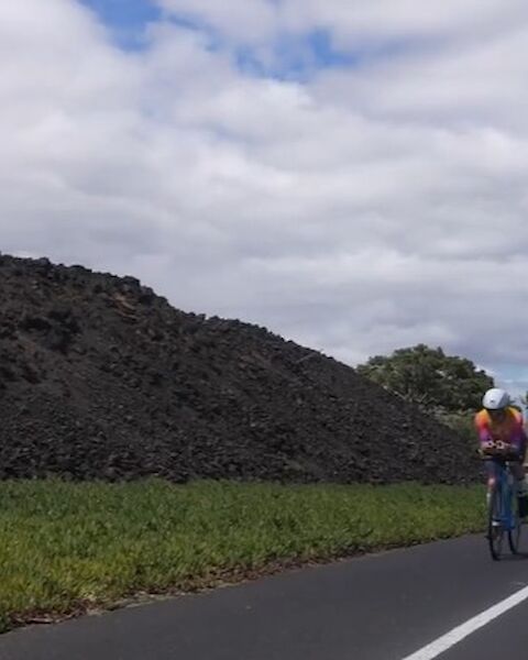 Two cyclists ride on a paved path beside a stone wall and green fields under a partly cloudy sky, with a few trees in the distance.