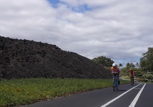 Two cyclists ride on a paved path beside a stone wall and green fields under a partly cloudy sky, with a few trees in the distance.