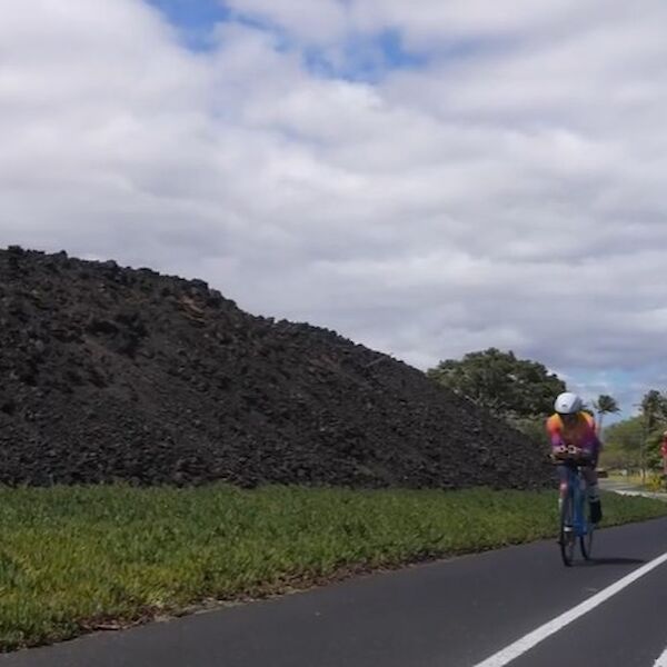 Two cyclists ride on a paved path beside a stone wall and green fields under a partly cloudy sky, with a few trees in the distance.