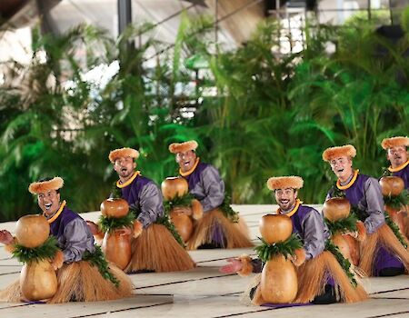 A group of small panda-like dolls wearing purple tops and straw skirts dance on a tiled surface with greenery in the background, holding orange fruit, all facing forward.