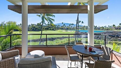 A sunny patio with wicker chairs, a table, and a view of a lush green golf course and palm trees beyond the glass railing, under a shaded structure.