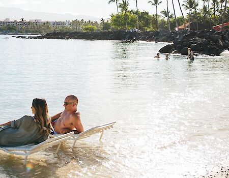 Two people relaxing on a paddleboard at a sunny beach, with calm water and a sandy shore in the background.