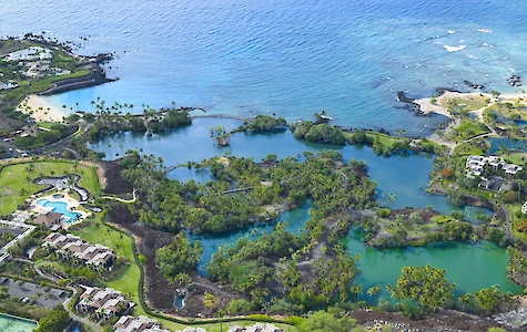 Tropical coast with emerald lagoons, lush trees, rocky shores, and luxury resort buildings along a bright blue ocean shoreline, sunny day.